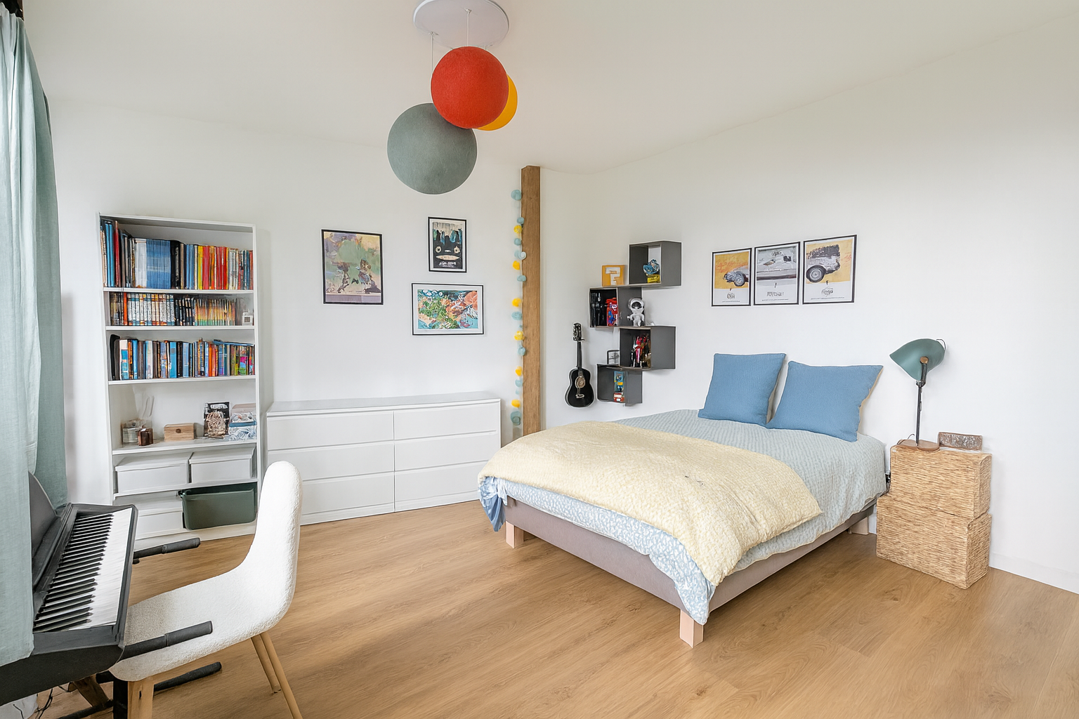 Une chambre avec un lit, parure de lit jaune et bleu à motif, coffres en osier, un lampadaire céladon, des étagères murales, une guitare suspendue, une bibliothèque blanche remplie de livres, et des décorations murales colorées.