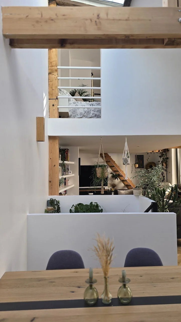 Intérieur d'une maison avec escalier en bois, plantes d'intérieur, livres, et une table avec des décorations en verre au premier plan.