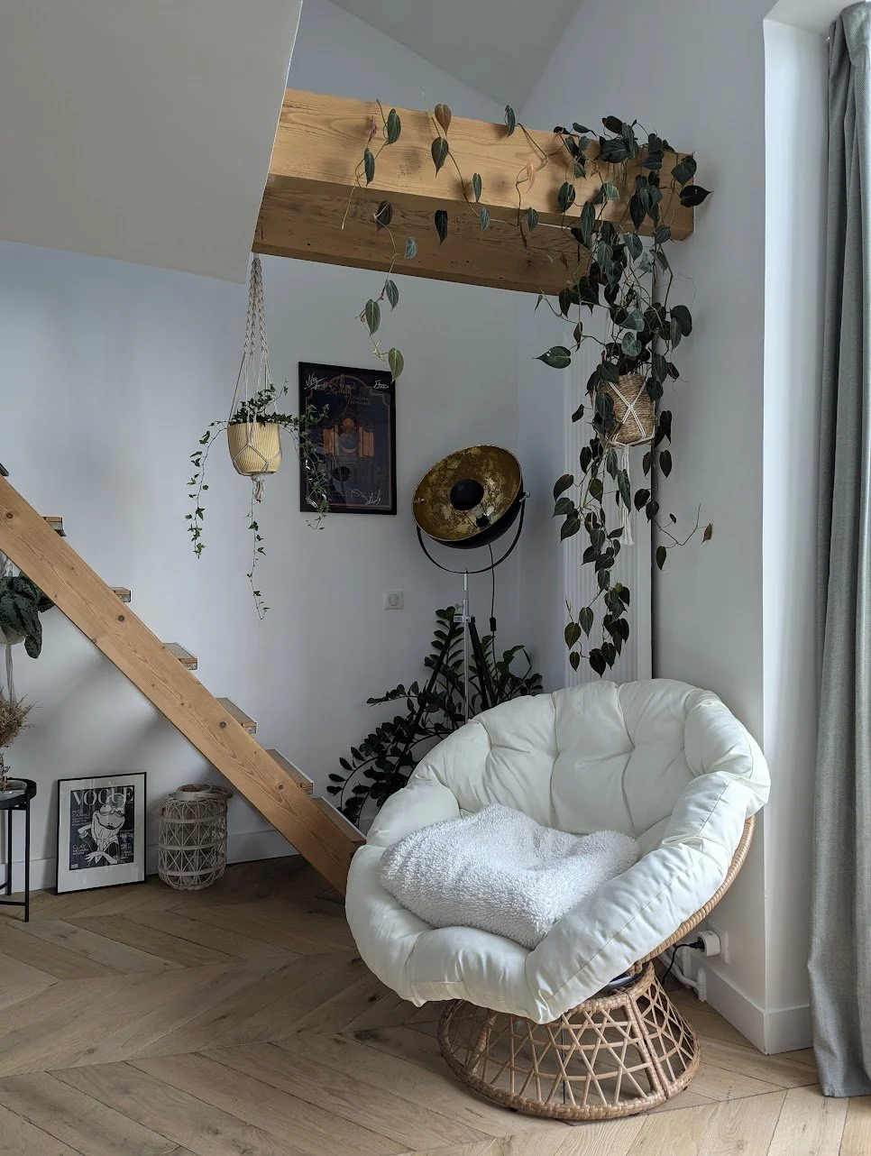 Coin salon avec fauteuil rond en rotin, coussin blanc, plantes en suspension et au sol, escalier en bois, lampadaire avec plafonnier doré, fenêtres avec rideaux gris.