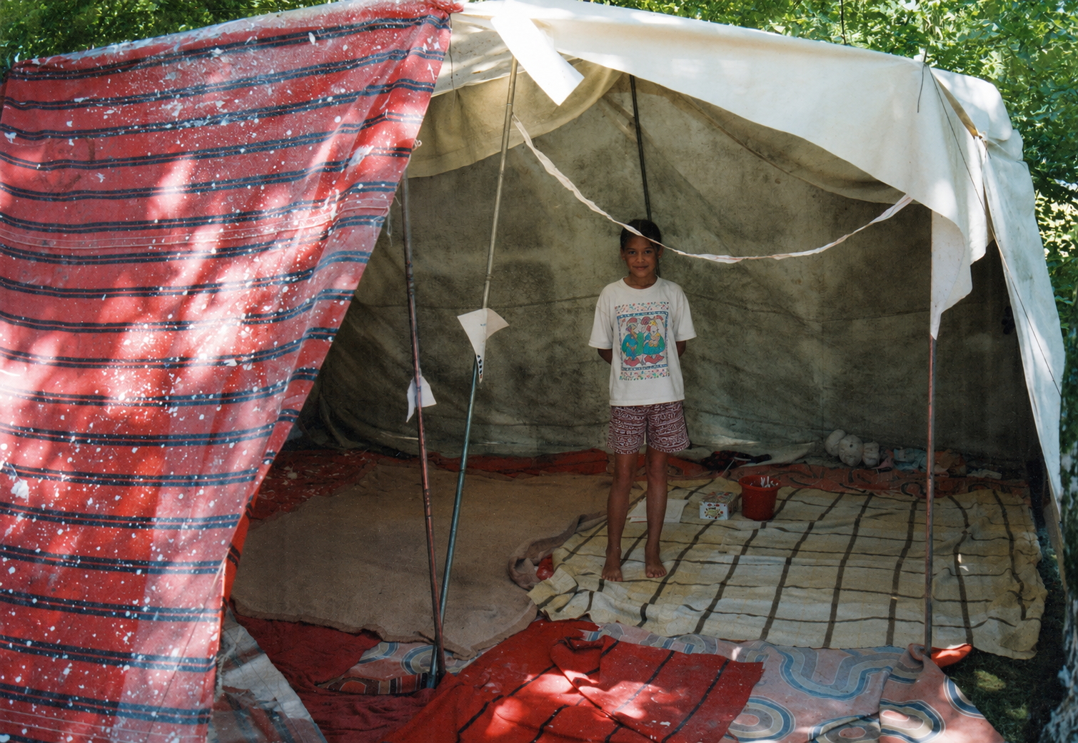 Une jeune fille souriante se tient à l'intérieur d'une tente en tissu ou en bâche, sous un arbre, avec un tapis et des objets en arrière-plan.