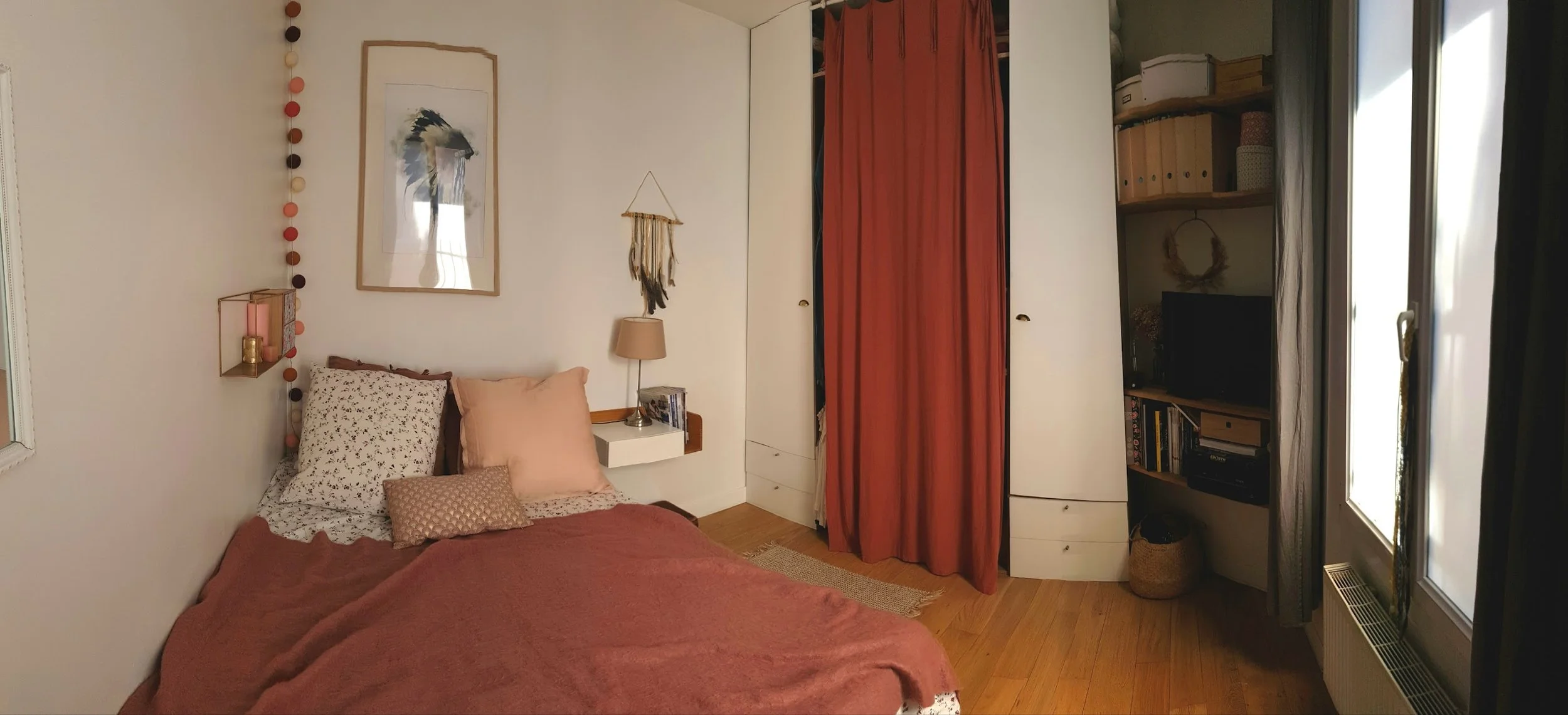 Chambre à coucher avec un lit, des oreillers, une étagère avec décorations, un meuble avec des livres, une télévision, et une fenêtre avec rideaux, décorée dans un style cosy.