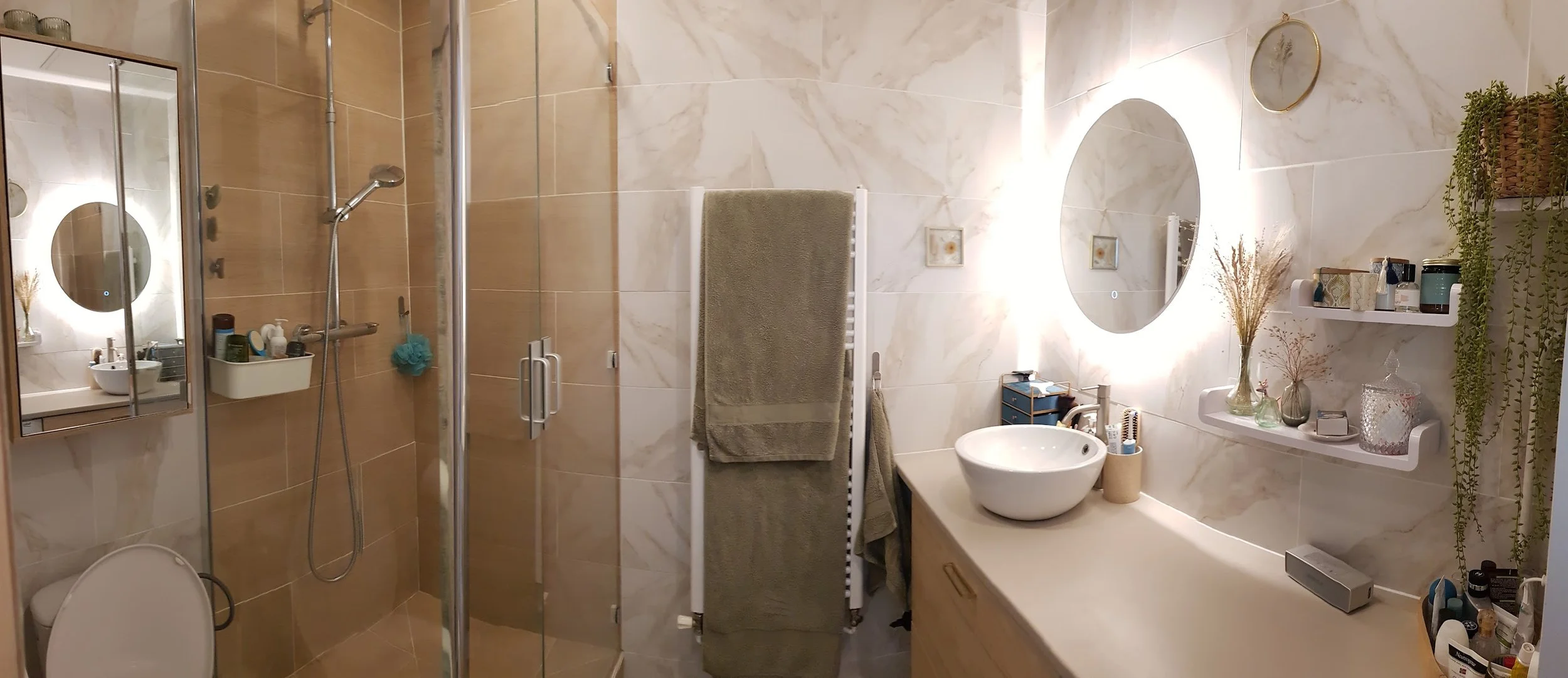 Salle de bain carrelage marbre & bois avec douche, vasque ronde à poser, miroir rond rétro éclairé, étagères blanches adhésives, plantes. draps de bain vert sauge
