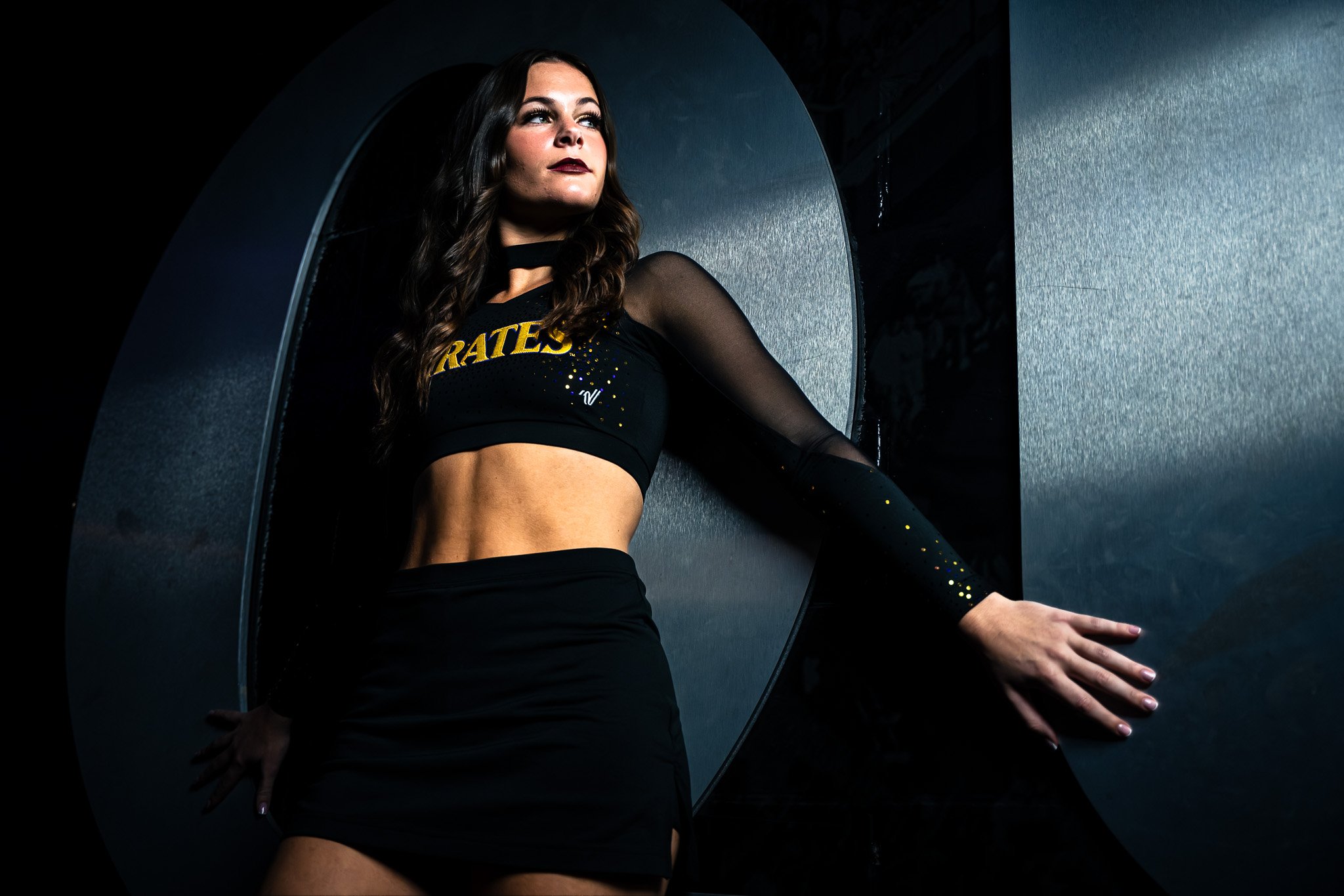 A woman with long dark hair and dark lipstick leaning against a reflective black surface with her hand on it, wearing a black sports bra with gold lettering and mesh sleeves.