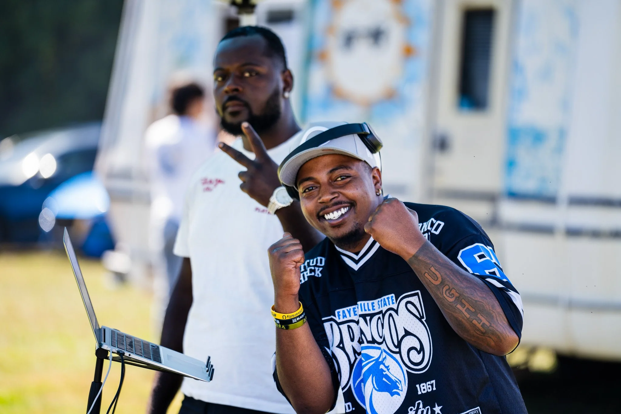 Two young men at an outdoor event, one smiling and posing with a fist near his chin, the other making a peace sign, stands behind a laptop on a stand, with a blurred crowd in the background.