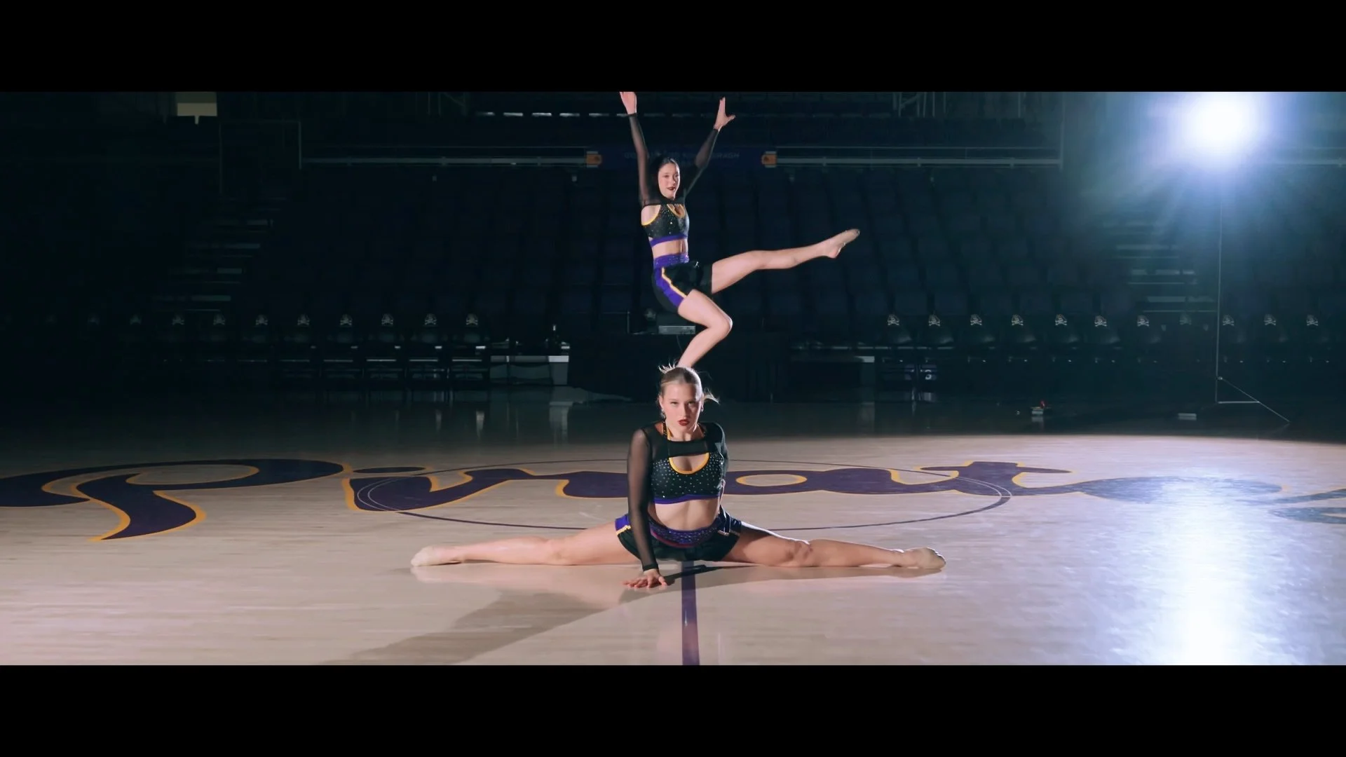 Three female gymnasts performing a routine on an indoor basketball court. One is in a split on the floor, another balancing on her hands with legs extended, and the third in the air above them.