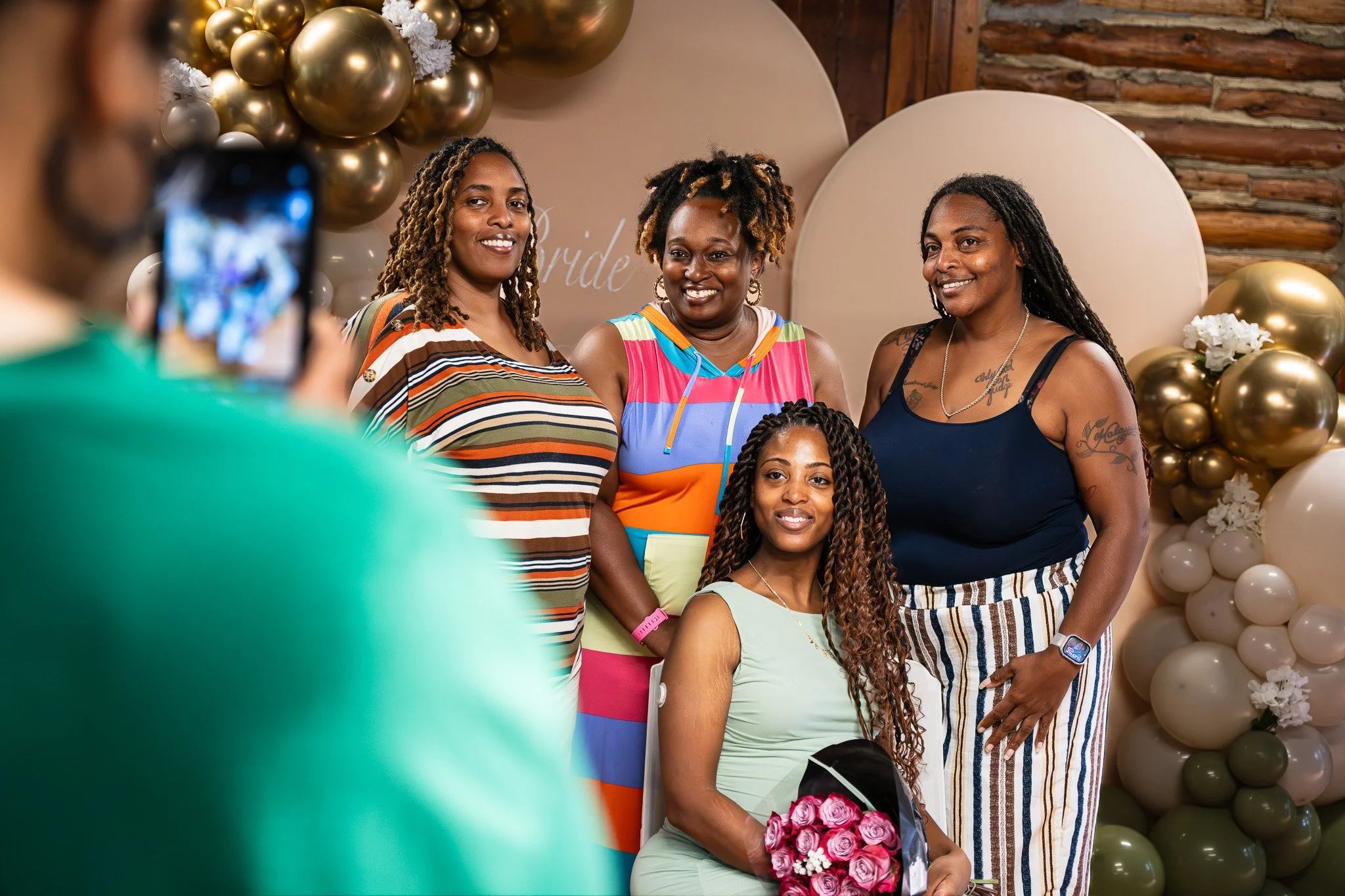 A group of women posing for a photo at a celebration or wedding event, with decorative balloons and flowers in the background.