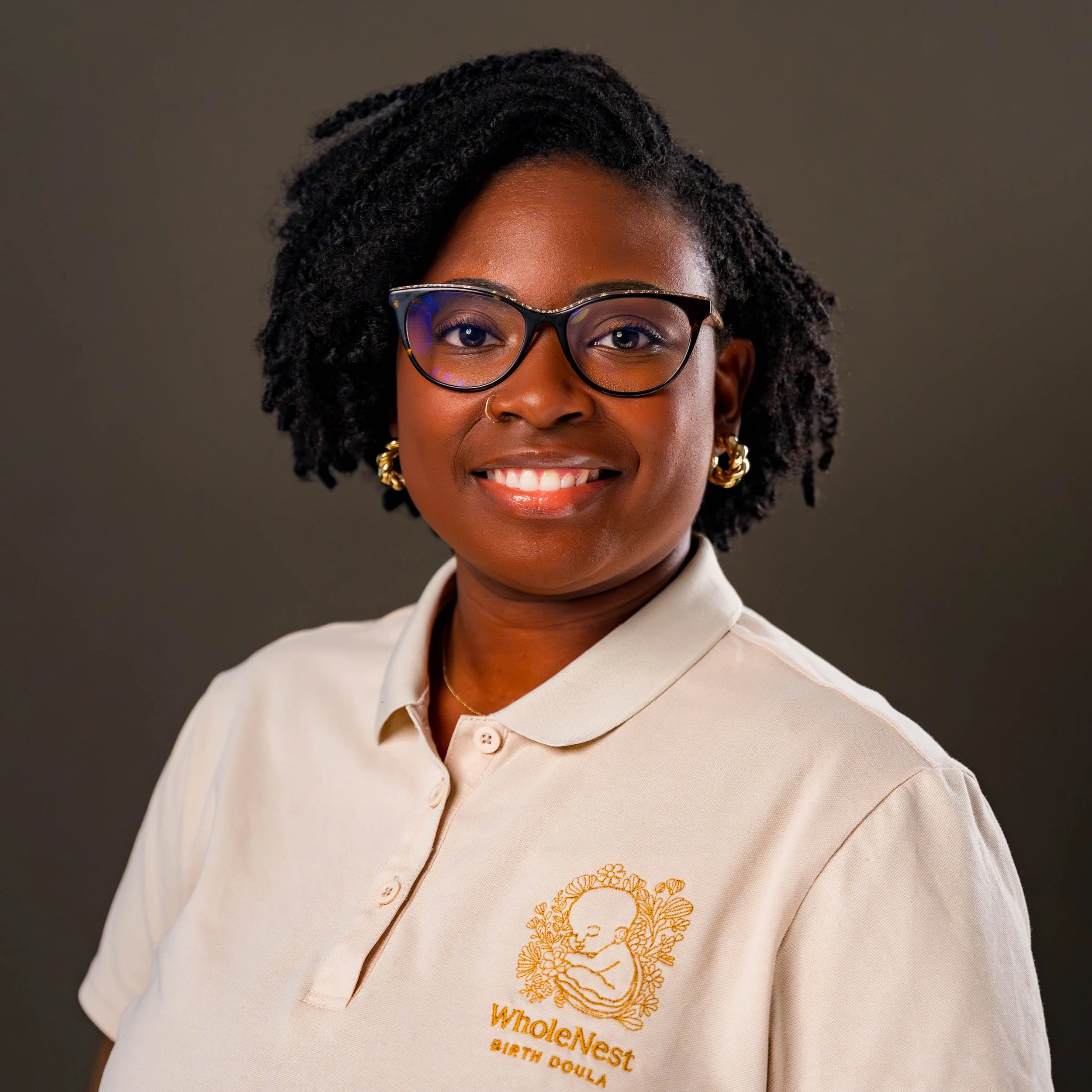 Close-up of a woman smiling, wearing glasses and earrings, with short, curly hair, dressed in a light-colored shirt with embroidered orange text and graphic on it.