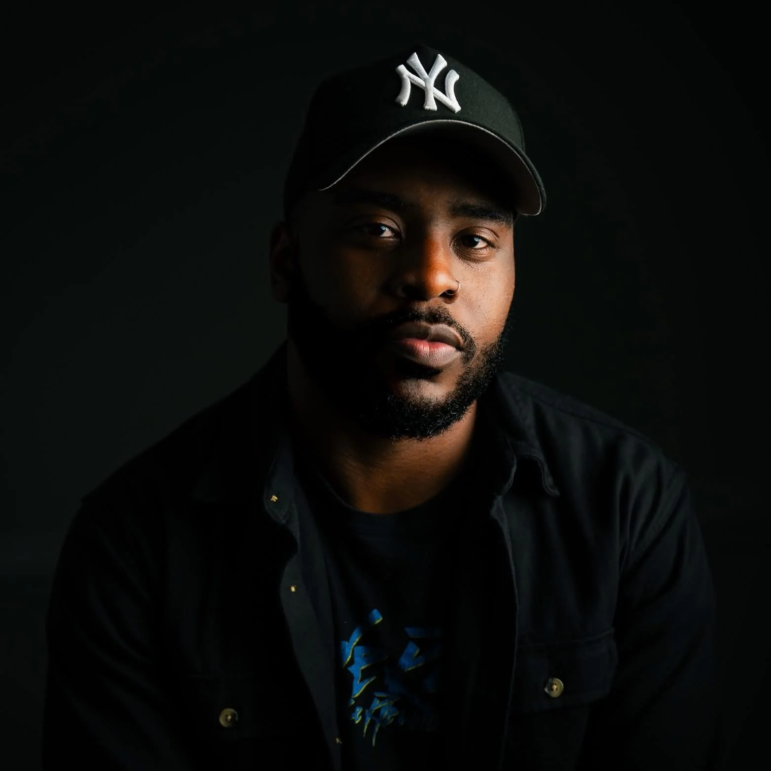 A portrait of a man wearing a black cap with New York Yankees logo and a black shirt, against a dark background.