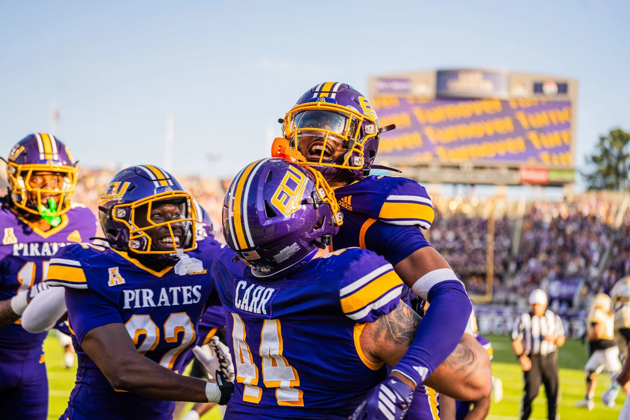 American football players in purple and yellow uniforms celebrating on the field during a game, with a crowd and a scoreboard in the background.