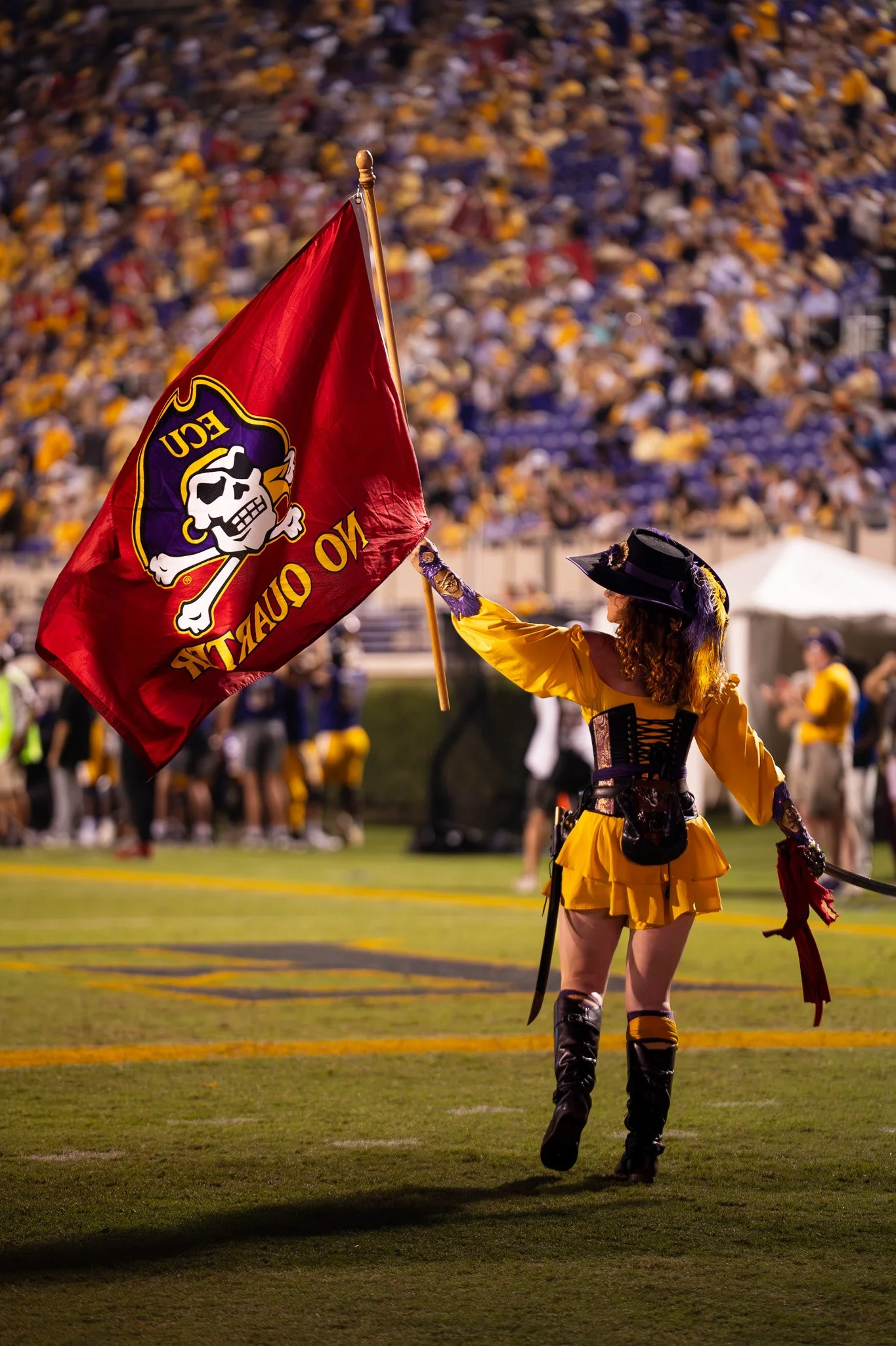 A person dressed as a pirate in a yellow outfit and black boots is holding a red pirate flag with a skull and crossbones, standing on a football field during an event with a large crowd in the background.