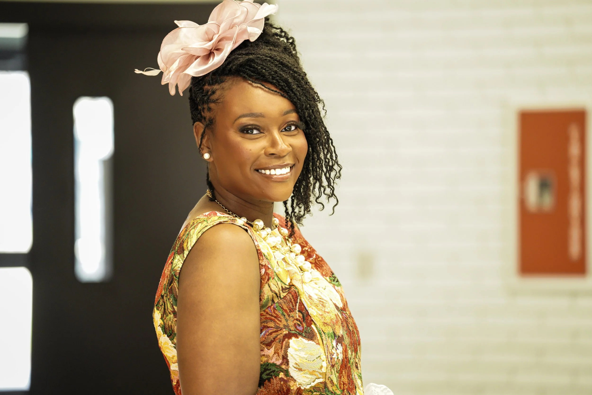 A smiling Black woman in a colorful, patterned dress and large pink headpiece, posing indoors with a background of a white wall and a door.