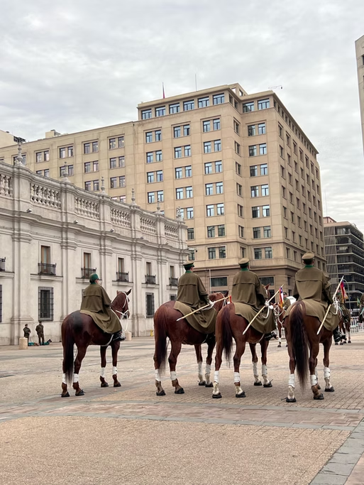 Quatro cavaleiros uniformizados em fileira, de espadas na mão, em uma praça com edifícios ao fundo