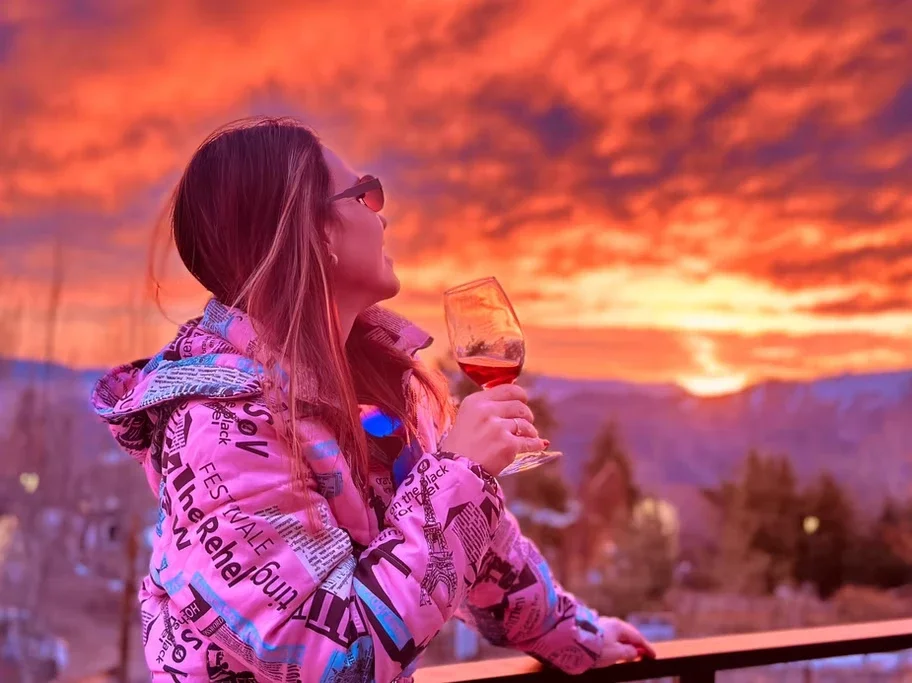 Mulher com óculos escuros segurando uma taça de vinho rosé, apreciando o pôr do sol alaranjado no céu, usando jaqueta com estampa de jornais.