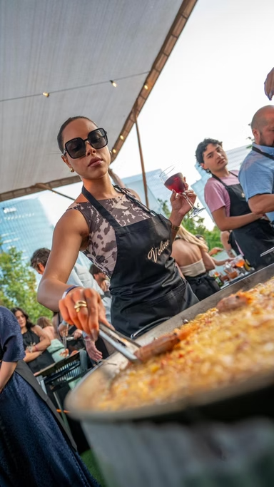 Mulher segurando um copo de vinho e usando um avental preto, preparando comida em uma festa ao ar livre.