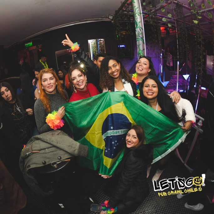Grupo de pessoas sorrindo segurando uma bandeira do Brasil em uma festa com clima festivo e luzes coloridas.