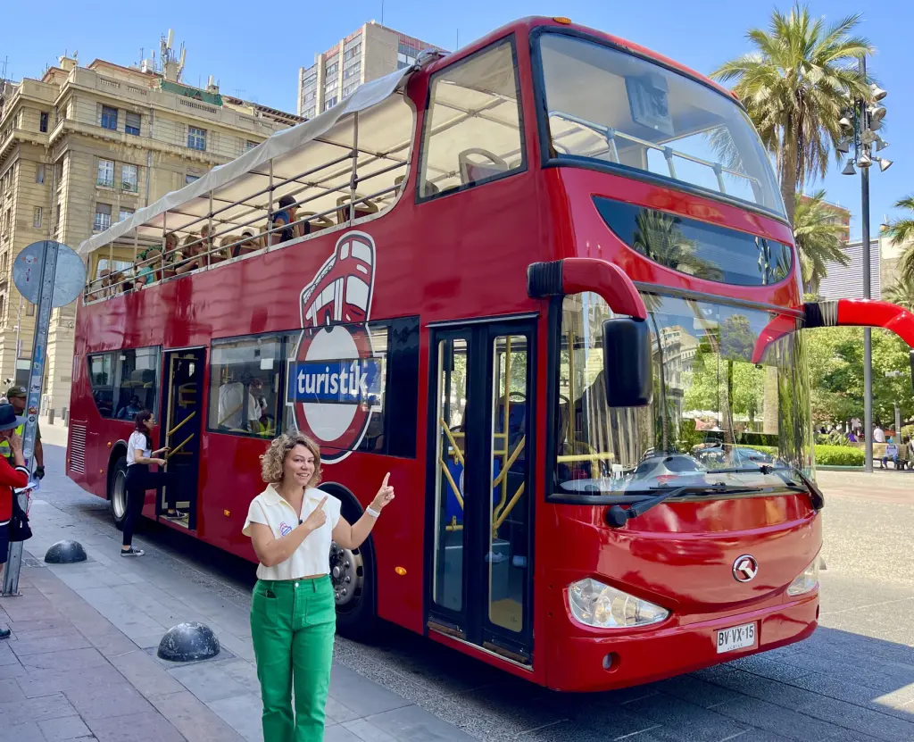 Um ônibus vermelho de dois andares, aberto na parte superior, com um logo de turismo e a palavra 'turistik'. Pessoas subindo e descendo do ônibus na rua, uma mulher sorrindo e apontando para o ônibus, em um dia ensolarado com árvores e edifícios ao fundo.
