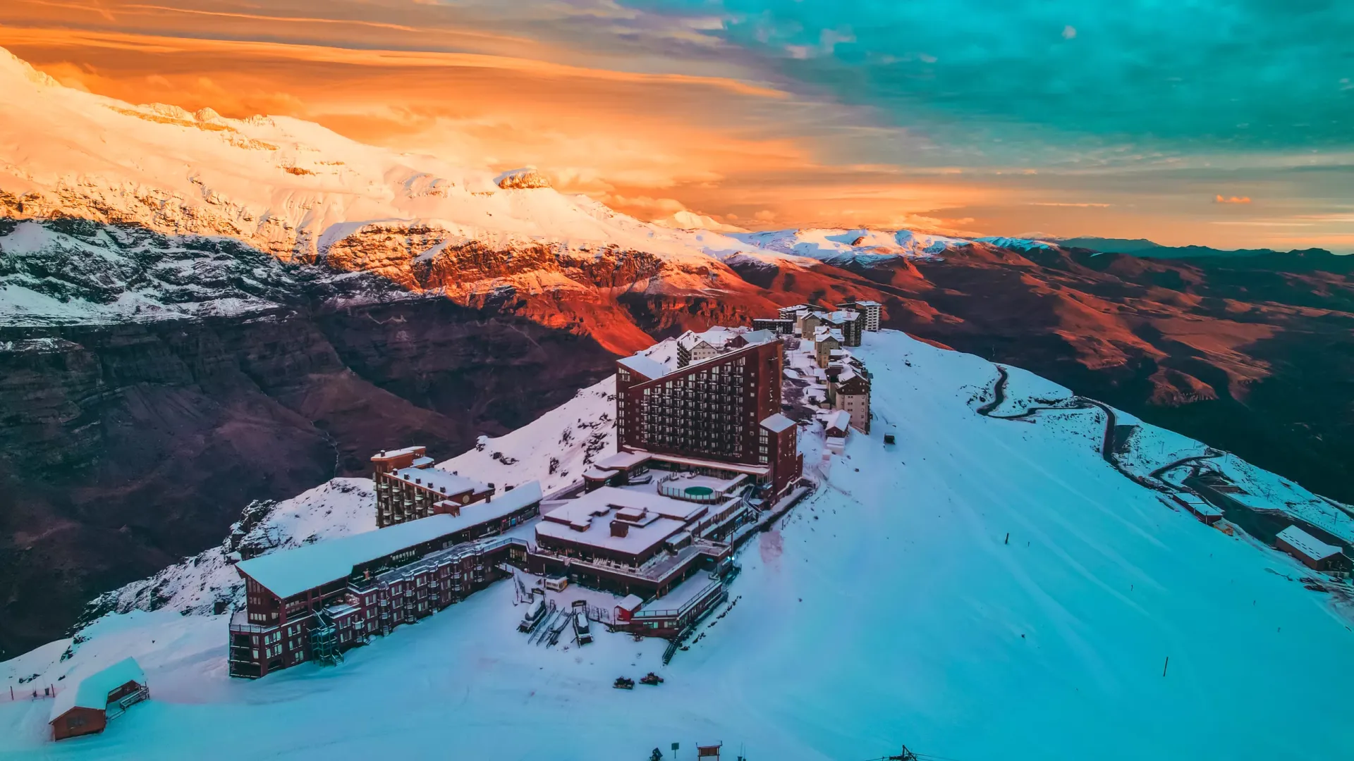 Vista panorâmica de uma cadeia de montanhas nevadas ao pôr do sol, com construções e um hotel no topo de uma colina coberta de neve.