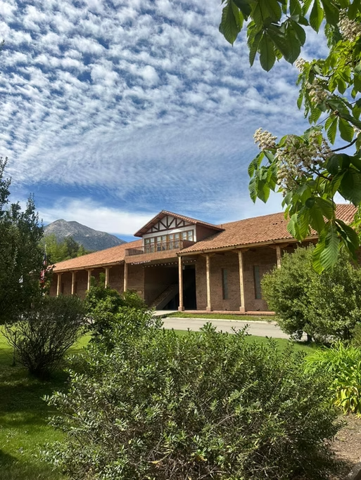 Casa de madeira com laje de telhado de cerâmica, rodeada de árvores verdes e arbustos, com montanhas ao fundo e céu com nuvens.