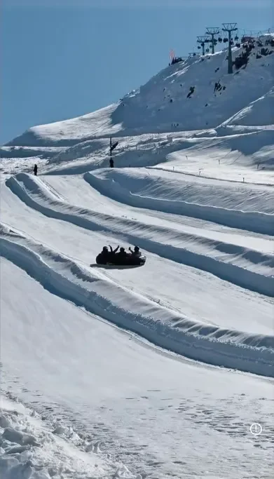 Pessoas deslizando em uma pista de trenó em uma estação de esqui coberta de neve, ao lado de uma montanha com cabos de teleférico.