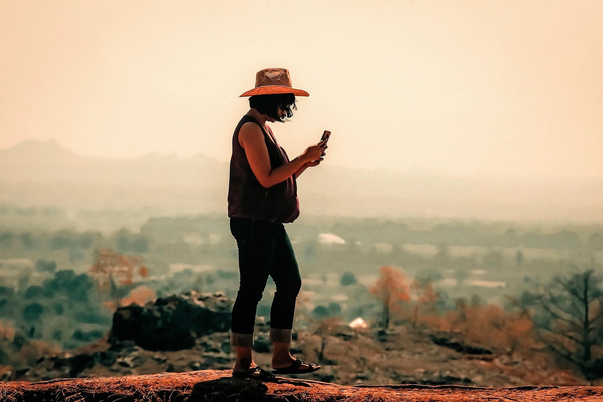 Pessoa mulher usando chapéu de palha, camiseta sem mangas, calça e chinelos, olhando para o celular em um ambiente ao ar livre durante o pôr do sol
