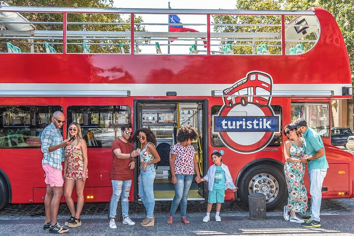 Grupo de pessoas ao lado de um ônibus de dois andares vermelho com o logo Turistik, no dia ensolarado.