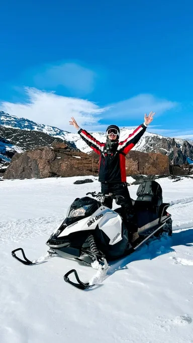 Pessoa com roupa de inverno e óculos de proteção em uma moto de neve em um cenário de neve, montanhas ao fundo, dia ensolarado.