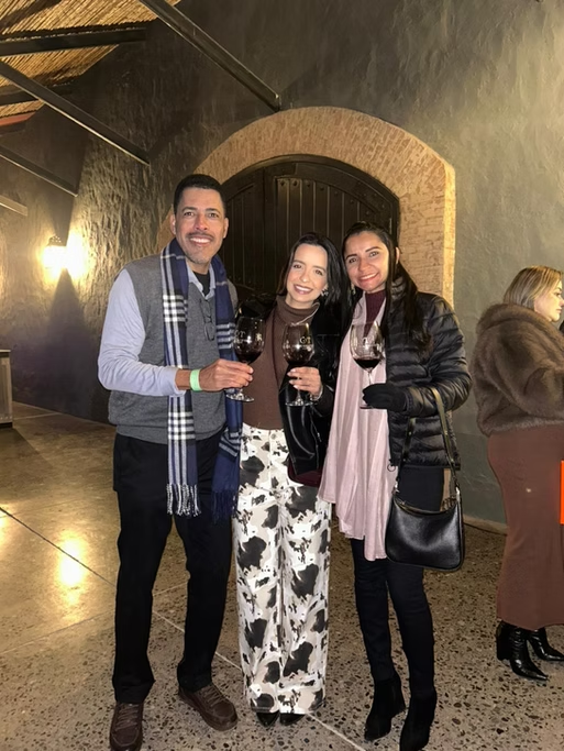 Três pessoas sorrindo segurando taças de vinho em um ambiente interno, com paredes de pedra, iluminação suave e uma mulher ao fundo vestindo casaco marrom.