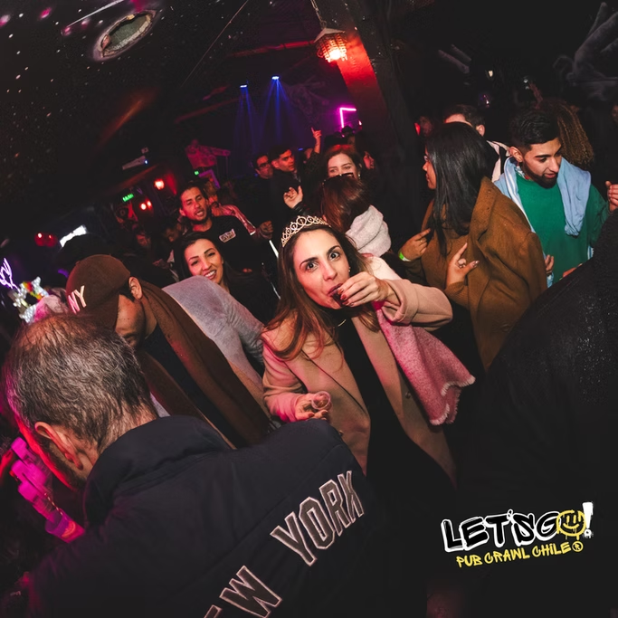 Pessoas em uma festa à noite, com iluminação de cores vibrantes, uma mulher usando coro e fazendo uma expressão divertida com a mão na boca, cercada por outros jovens dançando e socializando.