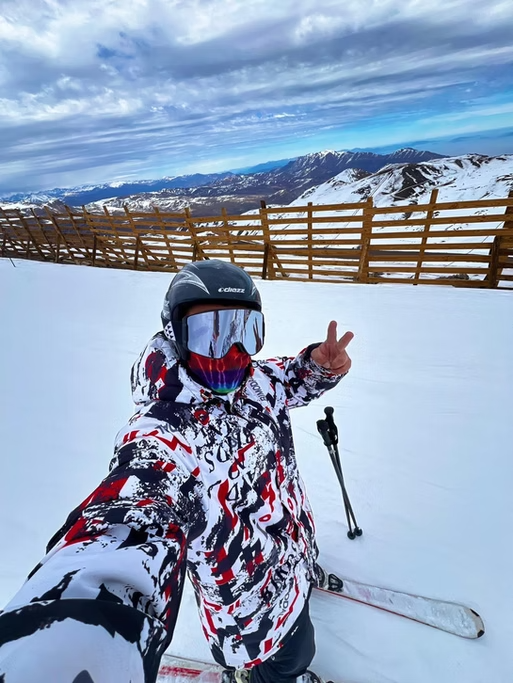 Pessoa vestido com roupas de esqui, usando capacete e óculos de proteção, segurando bastão de esqui, posando em uma paisagem de montanhas cobertas de neve com céu parcialmente nublado.