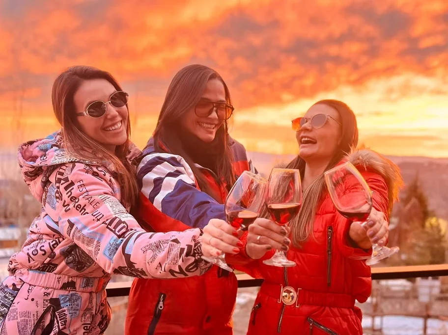 Três mulheres sorridentes brindando com taças de vinho tinto, com pôr do sol ao fundo.