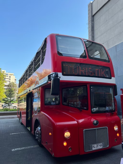 Duas andares de ônibus vermelho parado na rua, com painel digital mostrando como destino "Montik". Fundo com edifícios e céu claro.