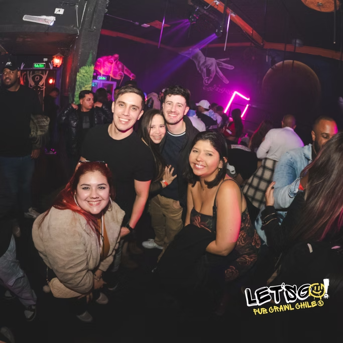 Grupo de amigos sorrindo em uma boate, com iluminação colorida e pessoas ao fundo.