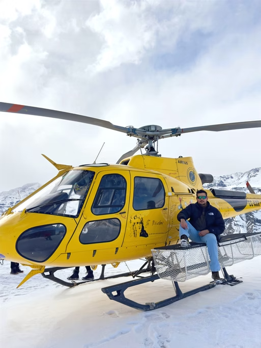 Homem com jaqueta azul e óculos escuros sentado na escada de uma aeronave de resgate amarela sobre neve com montanhas ao fundo.