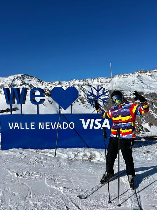 Pessoa com roupas de esqui segurando uma placa que diz "we ♥ VALLE NEVADO VIsa" em uma paisagem de montanhas cobertas de neve com céu azul.