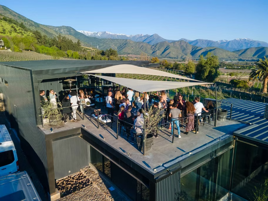 Pessoas reunidas em um terraço ao ar livre em um restaurante ou bar, com vista para montanhas ao fundo, sob céu claro e ensolarado.