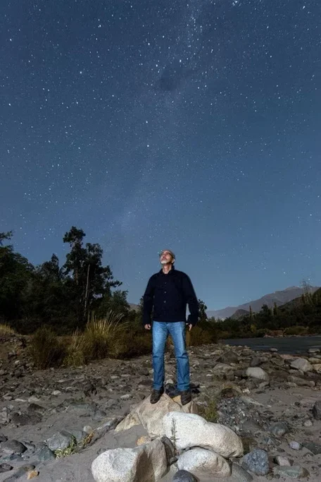 Homem de jaqueta escura e jeans observando o céu estrelado à noite, em um ambiente natural com árvores e montanhas ao fundo.