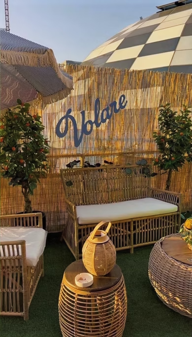 Área externa decorada com móveis de fibra natural, plantas, um toldo colorido, um mural de palha com a palavra 'Vollare' escrita em azul, e uma estrutura arquitetônica moderna ao fundo.