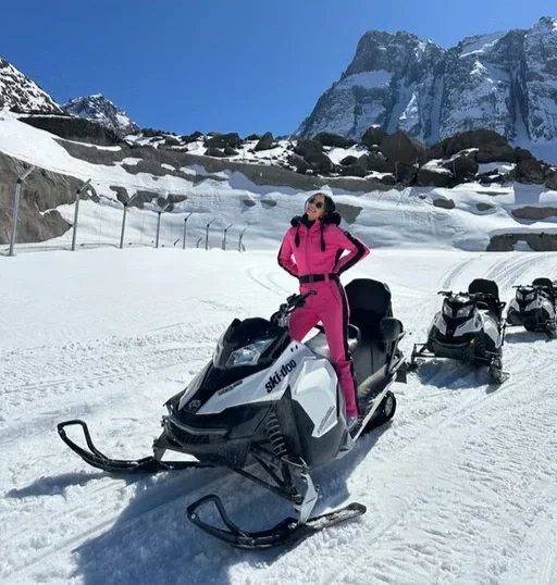 Pessoa de óculos e roupa rosa em uma moto de neve em uma paisagem de neve com montanhas ao fundo.
