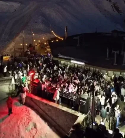 Festa ao ar livre com muitas pessoas, iluminação decorativa, e uma estrutura ao lado, possivelmente uma pista de dança ou palco.