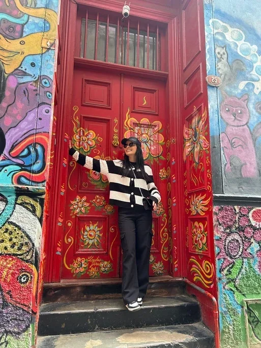 Pessoa vestida com casaco listrado preto e branco, calças pretas, óculos escuros e chapéu, de pé na porta vermelha decorada com desenhos florais, com graffiti colorido nas paredes ao redor.