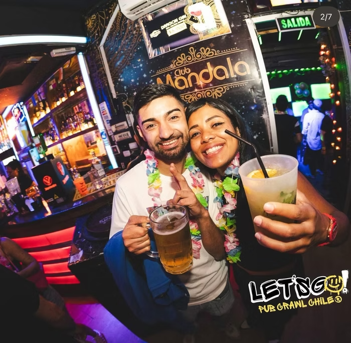 Dois amigos sorrindo e brindando com bebidas em um bar com decoração vibrante, iluminação colorida e uma placa de saída visível ao fundo na Clube Mandala, pub em Chile.