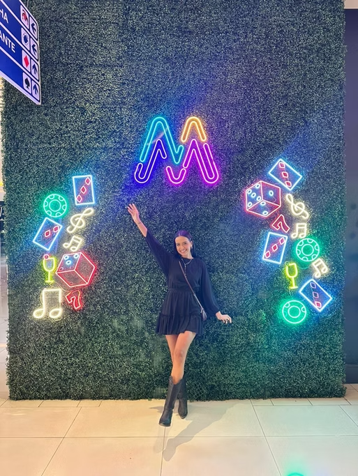 Mulher sorridente de vestido preto e botas pretas posando com braço levantado em frente a parede decorativa com neon colorido de símbolos de jogos e música, e uma logo de neon no topo central.