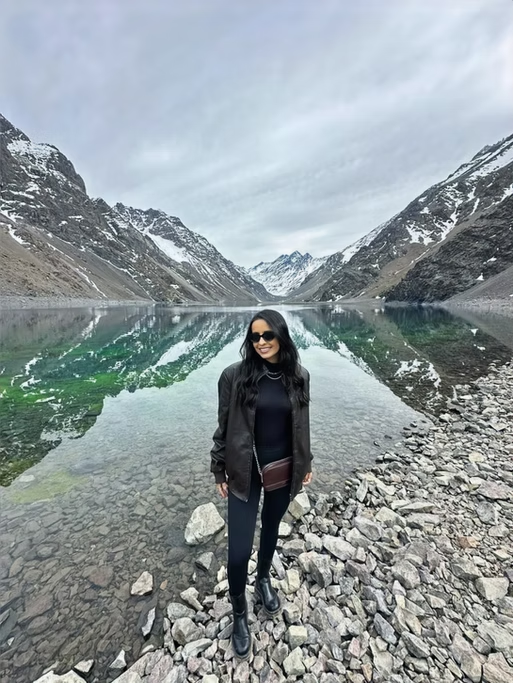 Mulher sorridente com óculos escuros, jaqueta preta, calça preta e botas, de pé na margem de um lago de montanha com águas verdes, cercado por picos cobertos de neve.