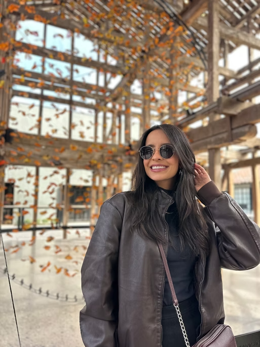 Mulher sorridente com óculos escuros, usando jaqueta de couro, em local com estrutura de madeira e folhas de outono caindo.