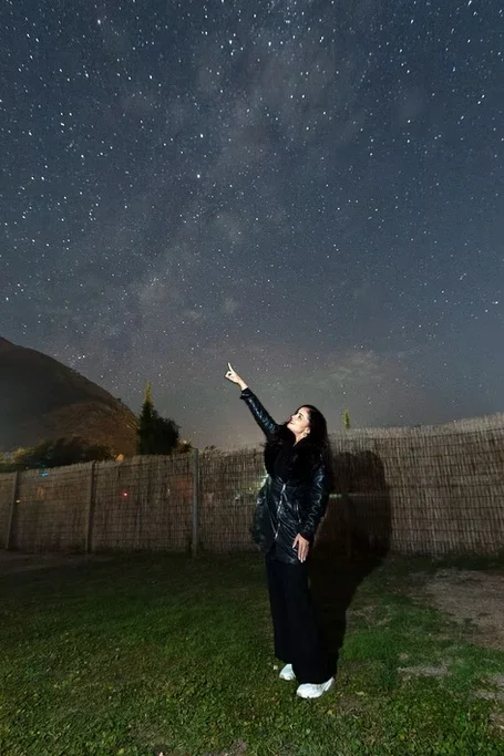 Mulher apontando para o céu estrelado ao ar livre à noite com uma cerca e montanhas ao fundo.