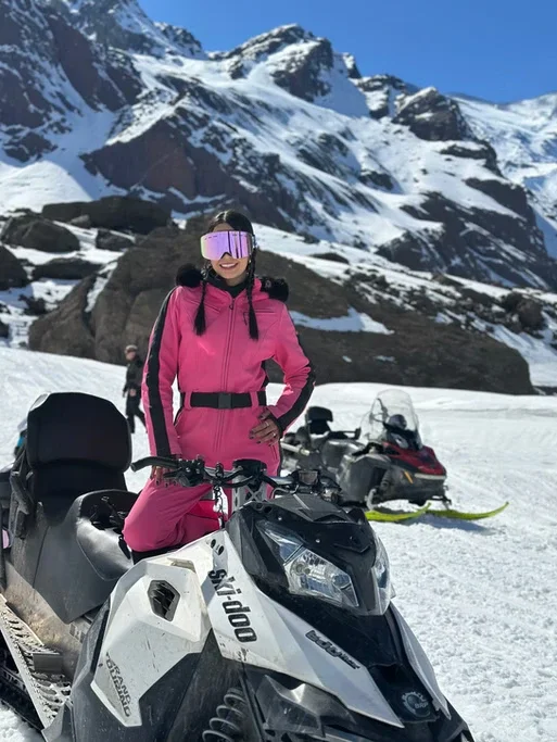 Mulher na neve, vestindo roupa rosa, com óculos de proteção, ao lado de uma moto de neve, com montanhas cobertas de neve ao fundo.
