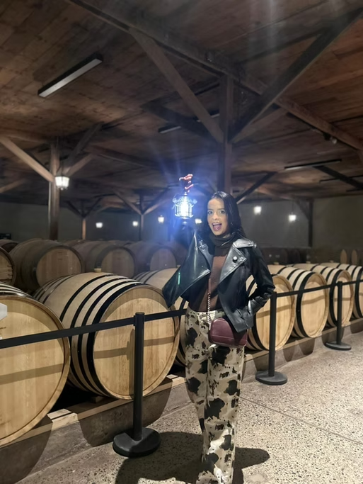 Mulher sorrindo em uma cave de vinhos, segurando uma lanterninha, com barris de vinho ao fundo.