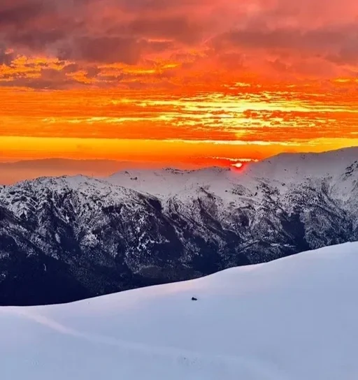Pôr do sol sobre uma cadeia de montanhas cobertas de neve, com céu em tons de laranja, vermelho e amarelo.
