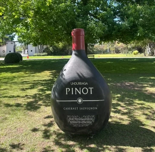Ampulheta gigante com rótulo de vinho, situada ao ar livre em um parque com árvores verdes ao fundo.