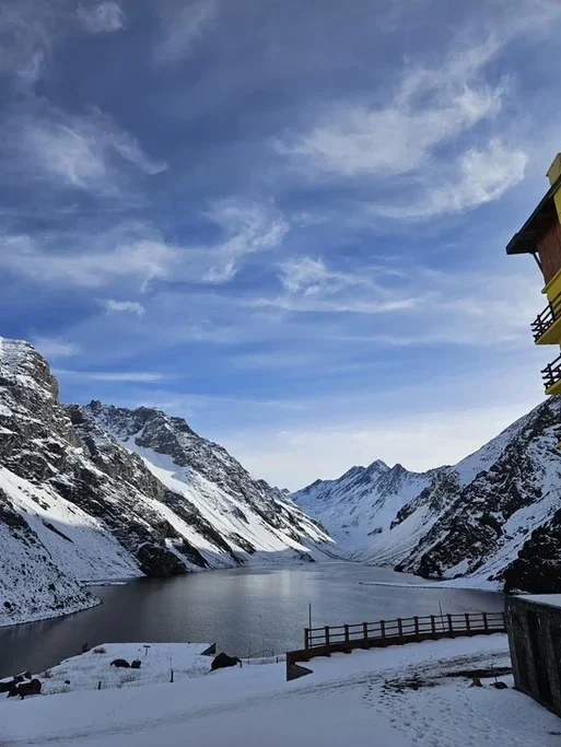 Vista de um lago em uma paisagem de montanhas cobertas de neve sob céu limpo com nuvens finas, ao lado de uma estrutura de edifício com varanda.
